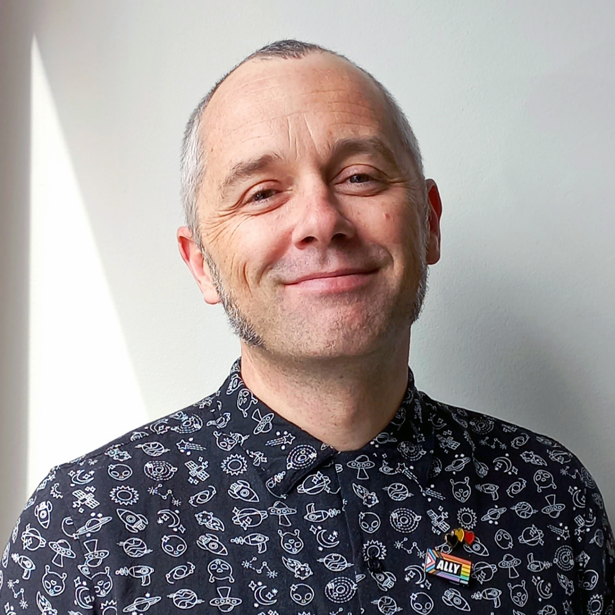 Man smiling in patterned shirt with ally pin against light wall background
